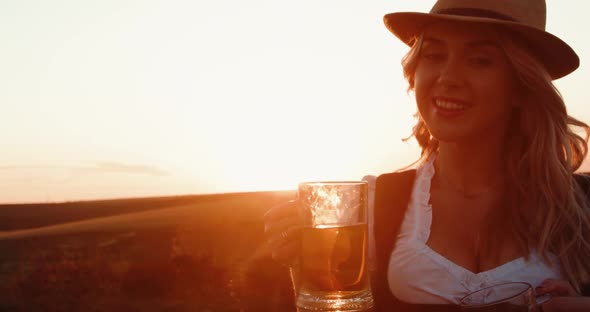 Girl in Hat and Dress Poses with Two Pints of Tasty Beer in Hands and Smiles