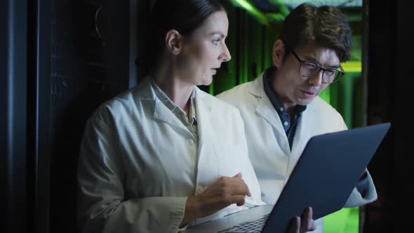 Diverse female and male it technicians in lab coats using laptop checking computer server