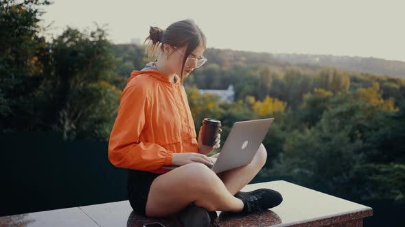 The Young Redhaired Woman with Glasses and Orange Blouse Taps Focused on the Keys of the Laptop