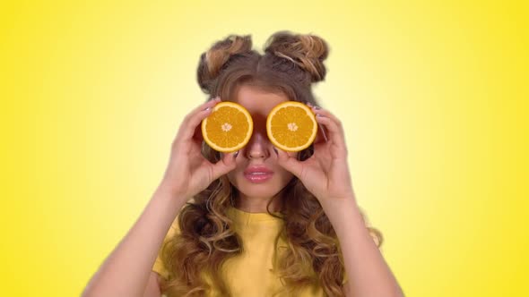 Attractive Girl in a Yellow Vest Closes Her Eyes with Oranges and Smiles Looking Into the Camera