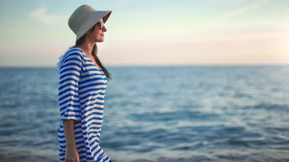 Happy Woman in Straw Hat and Sunglasses Walking on Seashore at Sunset