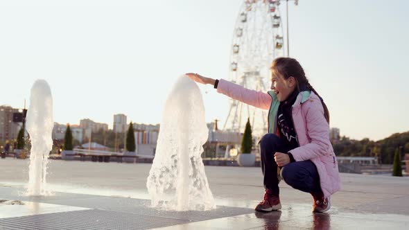 A Young Girl in a Pink Jacket Touches the Fountain with Her Hand
