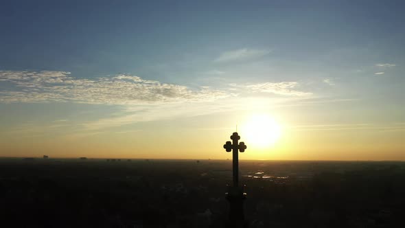 An aerial shot of a cathedral's steeple with a cross on top, taken at sunrise. The camera truck left