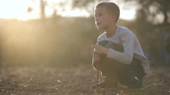 Child boy digging with a stick in the ground of black soil farm field at sunset