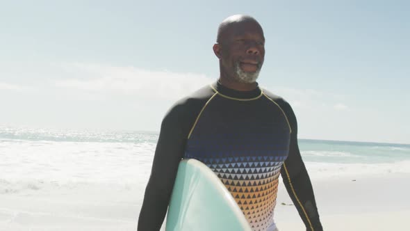 Senior african american man walking with surfboard on sunny beach