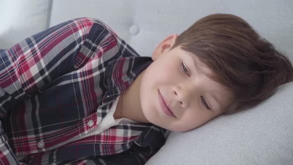 Close-up Portrait of Cute Caucasian Boy with Brown Hair and Grey Eyes Lying on Soft Pillow. Smiling