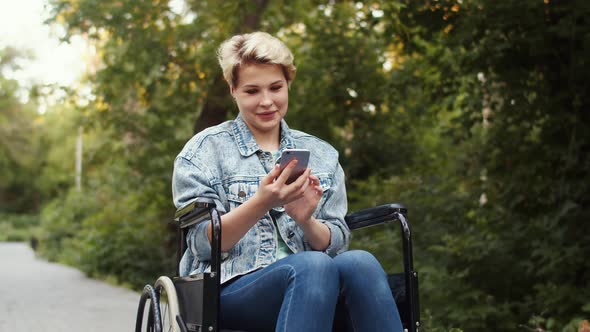 Young Disabled Woman is Sitting Wheelchair in Park and Chatting on Phone She Smiles and Types on