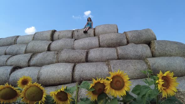 Young Girl Sitting on Top of the Round Hay Bale in the Sunflowers Field in Summer in Belarus