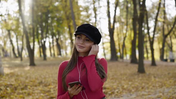 Glamorous Girl Posing in Autumn Park While Holding Phone and Pushing Earphones