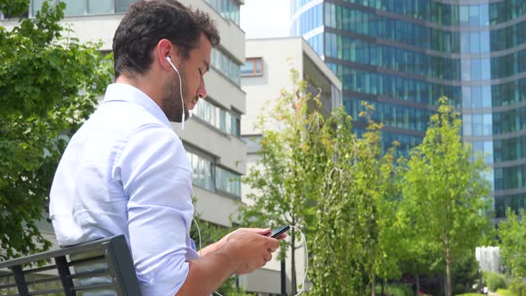 A Young Handsome Businessman Sits on a Bench, Puts on Earphones and Listens To Music