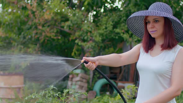 Young Female in White Dress and Elegant Hat Watering Vegetable Garden From Hose