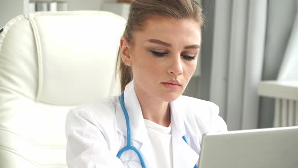 Portrait of Female Doctor in Medical White Coat and Stethoscope Works at Laptop in Clinic