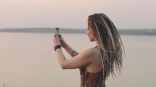 Attractive Hippie Woman with Dreadlocks Making Photo on Smart Phone at Sunset Near Sea