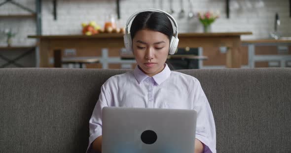 Asian Woman with Headphones Resting on Couch Using Laptop Computer Looking at Screen Typing Message