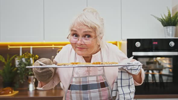 Grandmother in Apron is Smelling Hot Cookies on Baking Sheet and Smiling Being Satisfied Cooking at