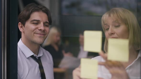 Positive Man and Woman Writing on Sticky Notes on Glass Door Talking Smiling