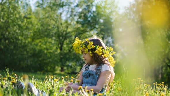 Little Cute Girl in a Flower Wreath on Her Head Sits on a Sunny Meadow Dreams Smiles and Looks