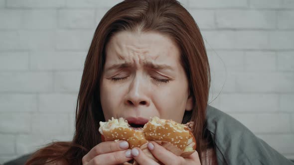 Unhappy Young Woman Eating Burger Looking At Camera In Bedroom