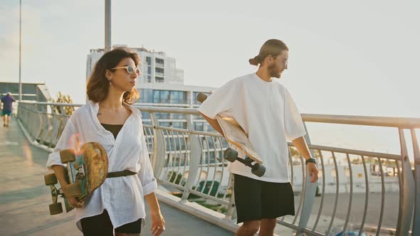 Young Man and Woman Going on Bridge with Skateboards Enjoying Sunset