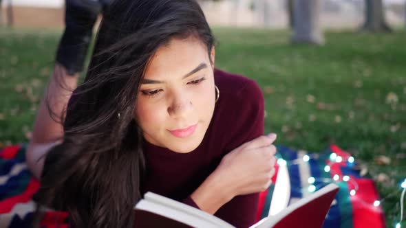 Close up on the face of a young hispanic woman reading the pages of a story book laying down outdoor