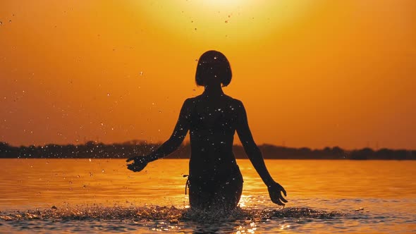 Silhouette of Woman at Sunset Raises Hands Up and Creating Splashes of Water