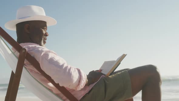 Smiling senior african american man lying on sunbed and reading book on sunny beach