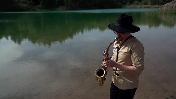 A Man with Glasses and a Hat Plays the Saxophone on a Summer Day on the Shore of a Lake, in Nature