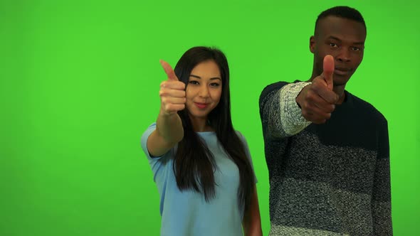 A Young Black Man and a Young Asian Woman Smile and Show Thumbs Up To Camera - Green Screen Studio