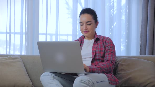 Young Serious Woman Freelancer Working on a Laptop Sitting on a Sofa at Home, Businesswoman Typing