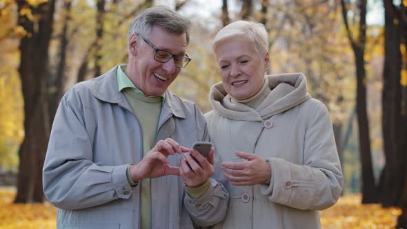 Joyful Caucasian Elderly Couple Walking in Autumn Park Hold Smartphone Use Mobile Internet Browser