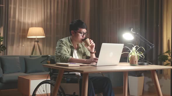 Asian Man Sitting In A Wheelchair While Working On Laptop Computer And Drinking Coffee At Home