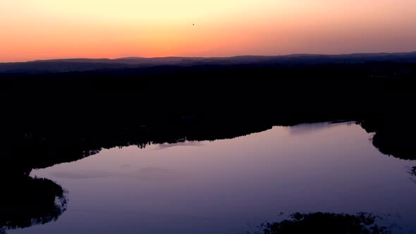 Aerial Over Calm Smooth Lake With Purple Reflected Light Against Orange Sunset Skies Silhouetted By