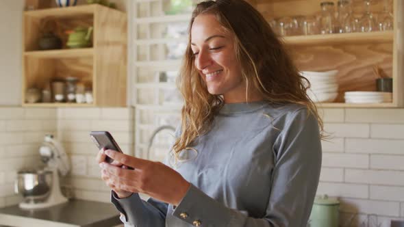 Happy caucasian woman standing in sunny cottage kitchen using smartphone and smiling