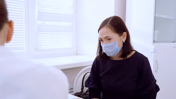 A Woman in a Medical Protective Mask Sits at a Table Against the Background of a Window