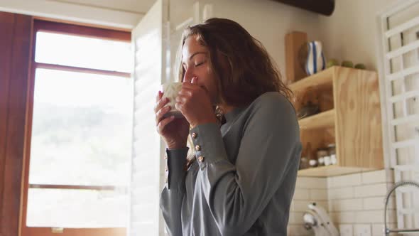 Happy caucasian woman standing in sunny cottage kitchen enjoying drinking coffee and smiling