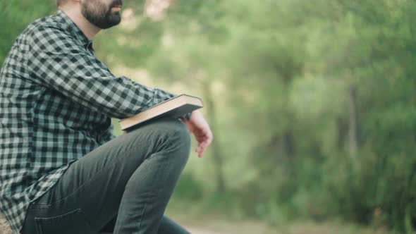 Caucasian Man With Book Sitting On The Ground With Green Nature In The Background. - close up