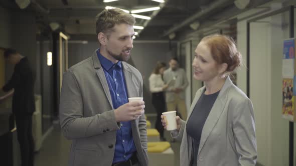 Two Caucasian Coworkers Talking in Open Space Office During Break. Man and Woman Holding Coffee Cups