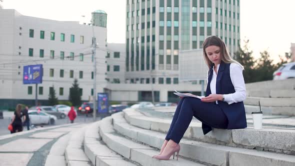 A young girl in an office suit in the city center sitting on the stairs, studying the paper and drin
