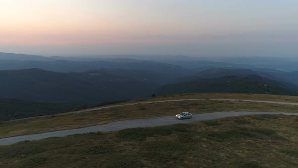 Aerial View of Car Driving on Sunset Mountain Road with Green Hills Valley and Dramatic Sky