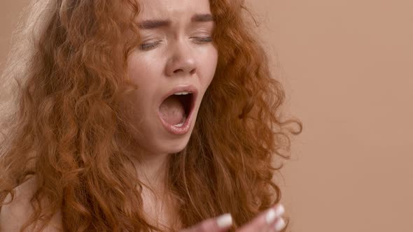 Sleepy RedHaired Young Woman Yawning Covering Mouth Beige Background