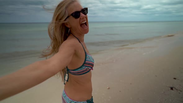 Slow motion with flying hair, a playful, mature woman in sunglasses and bikini running along the bea