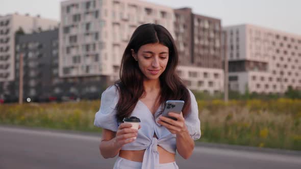 Beautiful Brunette Woman Holds Cell Phone and Cup of Coffee Uses Smartphone on the Street