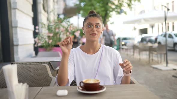 Girl is Sitting in Outdoor Coffee Shop and Speaks to the Camera