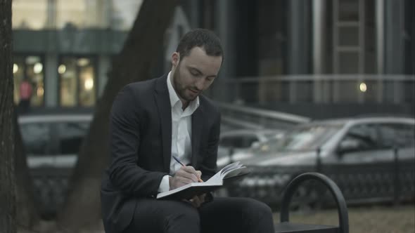 Young Bearded Business Man Thinking and Wrighting Notes at Green Park, Traffic Behind