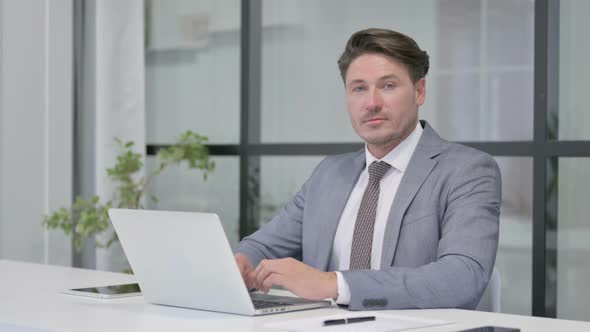 Middle Aged Man Smiling at Camera while using Laptop in Office