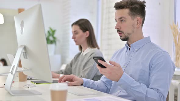 Focused Creative Man Using Smartphone and Working on Desktop