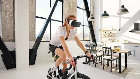 Young Female in VR Glasses is Pedaling on Indoor Exercise Bike Trainer