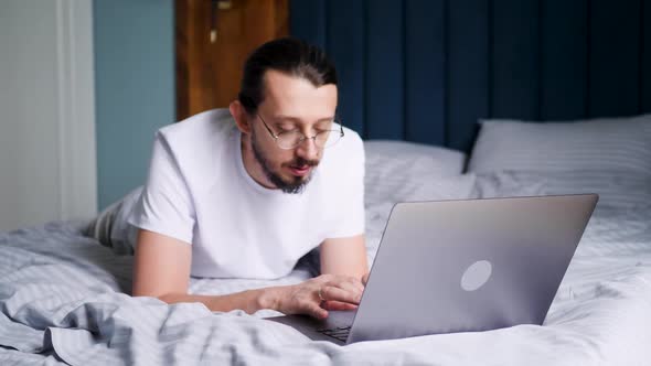 Funny Dance Bearded Man in Glasses and a Tshirt is a Freelancer Sitting on a Bed with a Laptop in an