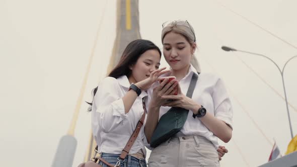 Asian lesbian couples using smartphones browsing the website while standing on the bridge.