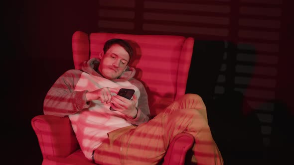 Young Man Browsing Smartphone Sitting in Armchair in Dark Room with Falling Red Light and Shadow